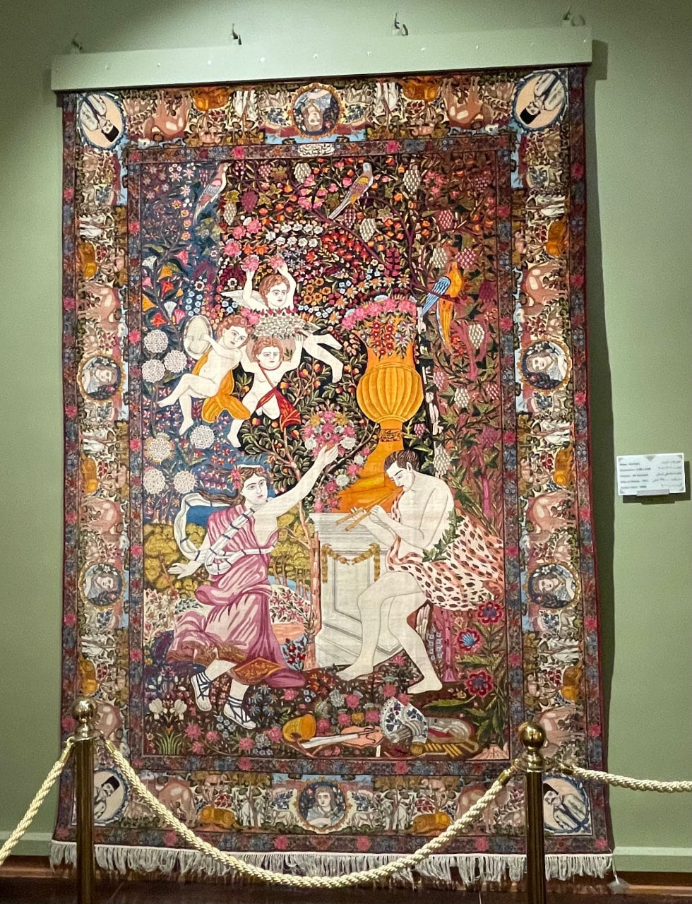 Photo of a large carpet draped in a museum, featuring a floral scene with dancing people